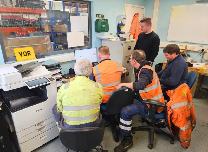 A group of five workers is gathered in a small office space, focusing on a computer screen. Four of the workers are seated, wearing high-visibility safety vests, and appear to be discussing something displayed on the monitor. One worker, standing behind them, is wearing a dark shirt and is engaged in the discussion. The office is equipped with various items including a printer, a whiteboard, and shelves with documents and supplies. Through a window, an industrial work area with equipment and tools is visible. The atmosphere suggests a collaborative session, possibly for planning or troubleshooting.