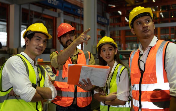 A group of four construction workers is seen standing together inside an industrial facility. They are wearing safety helmets and reflective vests. One worker, holding a folder with documents, is pointing towards something off-camera while explaining to the group. The others are attentively looking in the same direction, appearing engaged and focused on the explanation. The background features industrial equipment and structures, indicating an active work environment.
